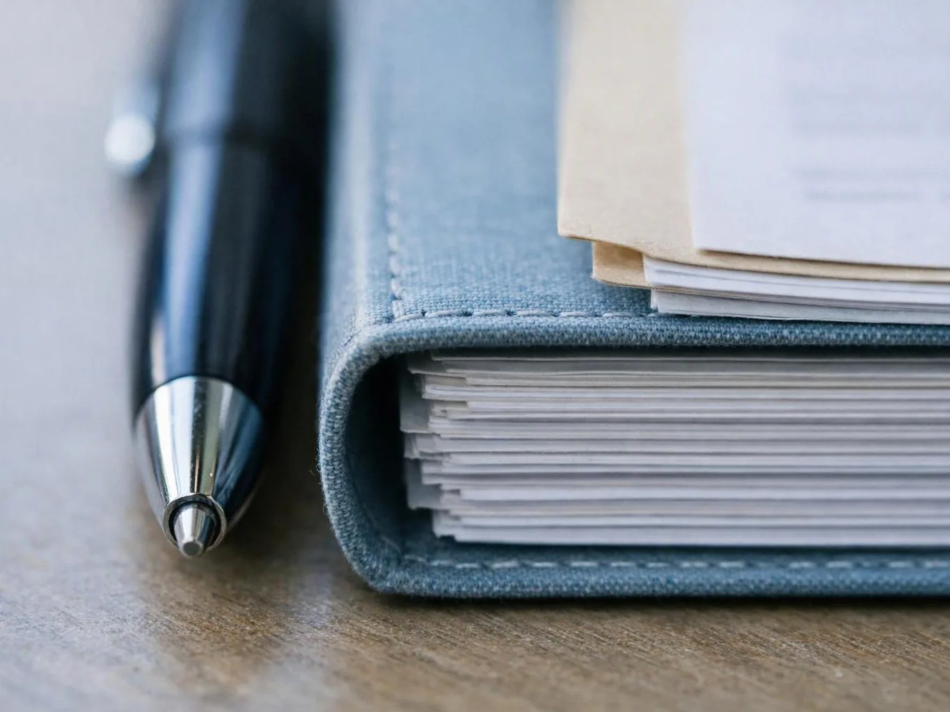 A black pen rests beside a blue fabric notebook stuffed with stacked papers on a wooden desk
