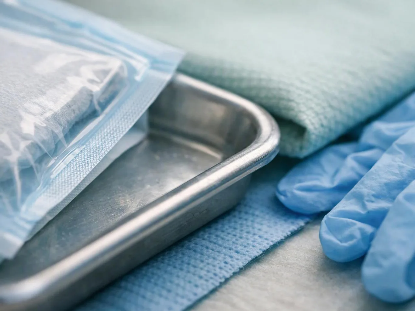 Blue medical gloves rest beside a stainless tray and sealed instrument pouch on a sterile surface