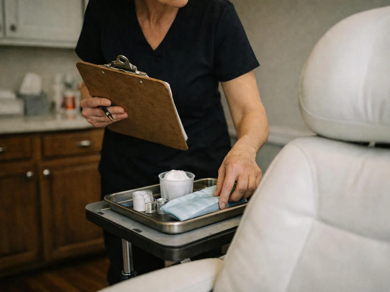 A clinician in dark scrubs checks a tray of medical supplies beside a padded exam chair