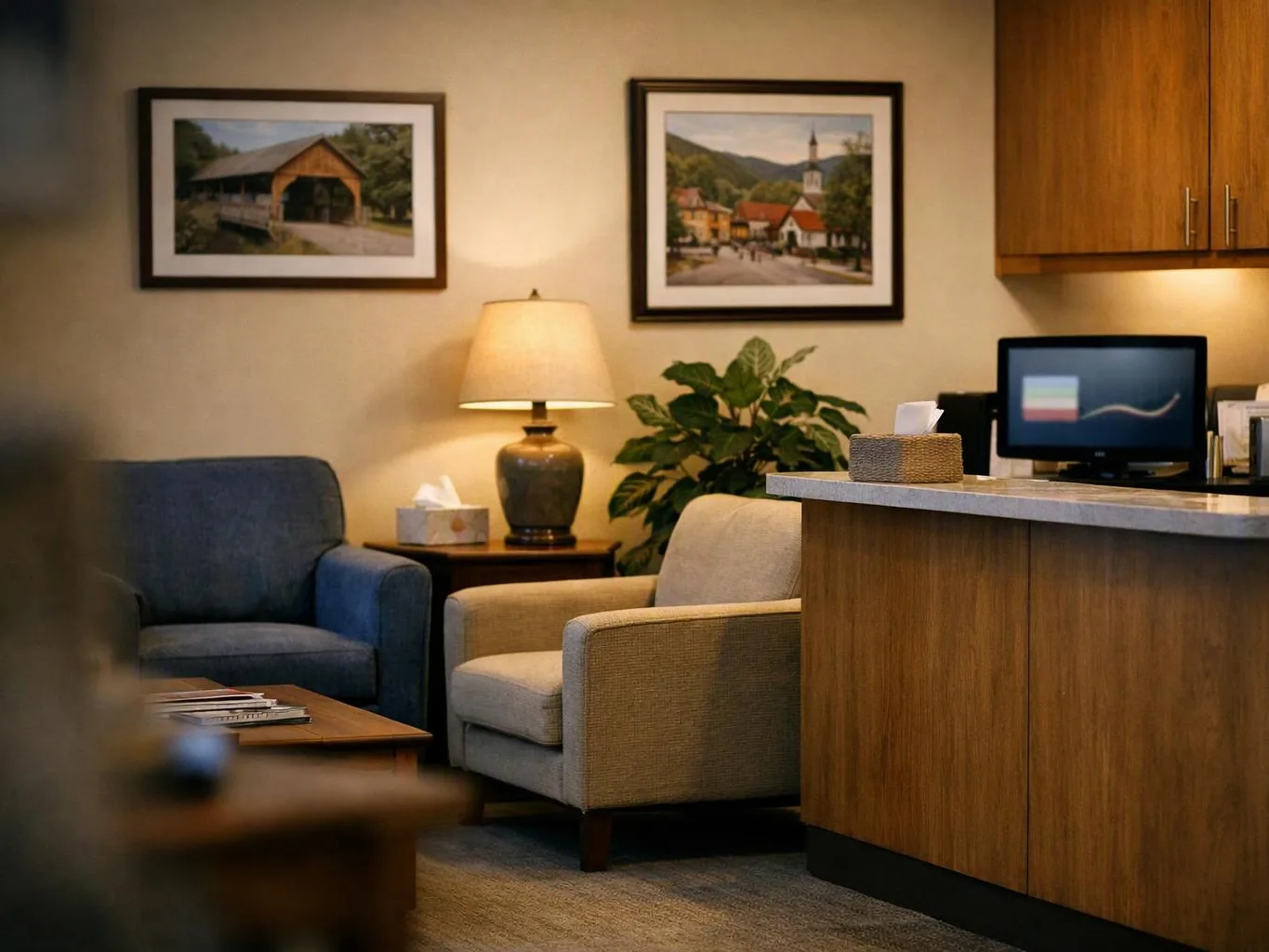 A cozy waiting room shows two chairs, a lamp, framed pictures, a plant, and a reception desk with a small monitor