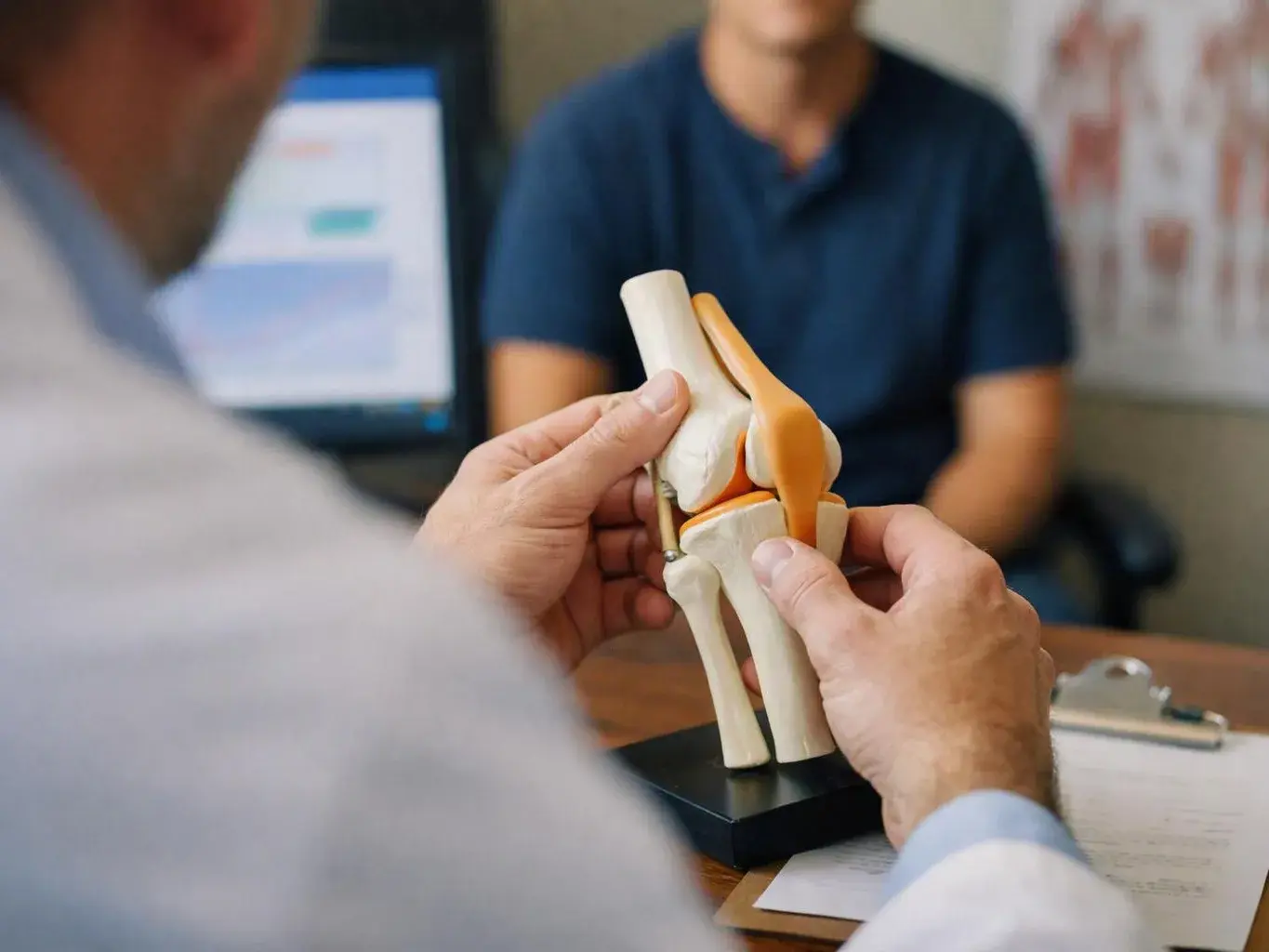 A doctor holds a knee joint model across a desk while speaking to a seated patient in an exam room