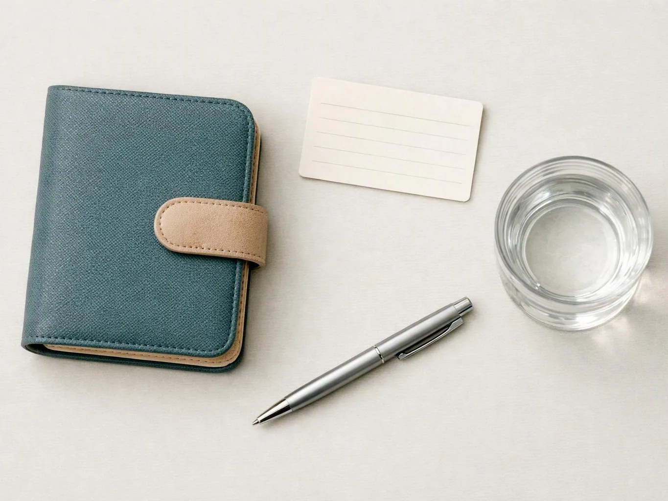 A teal notebook, blank card, silver pen, and clear glass sit on a white tabletop